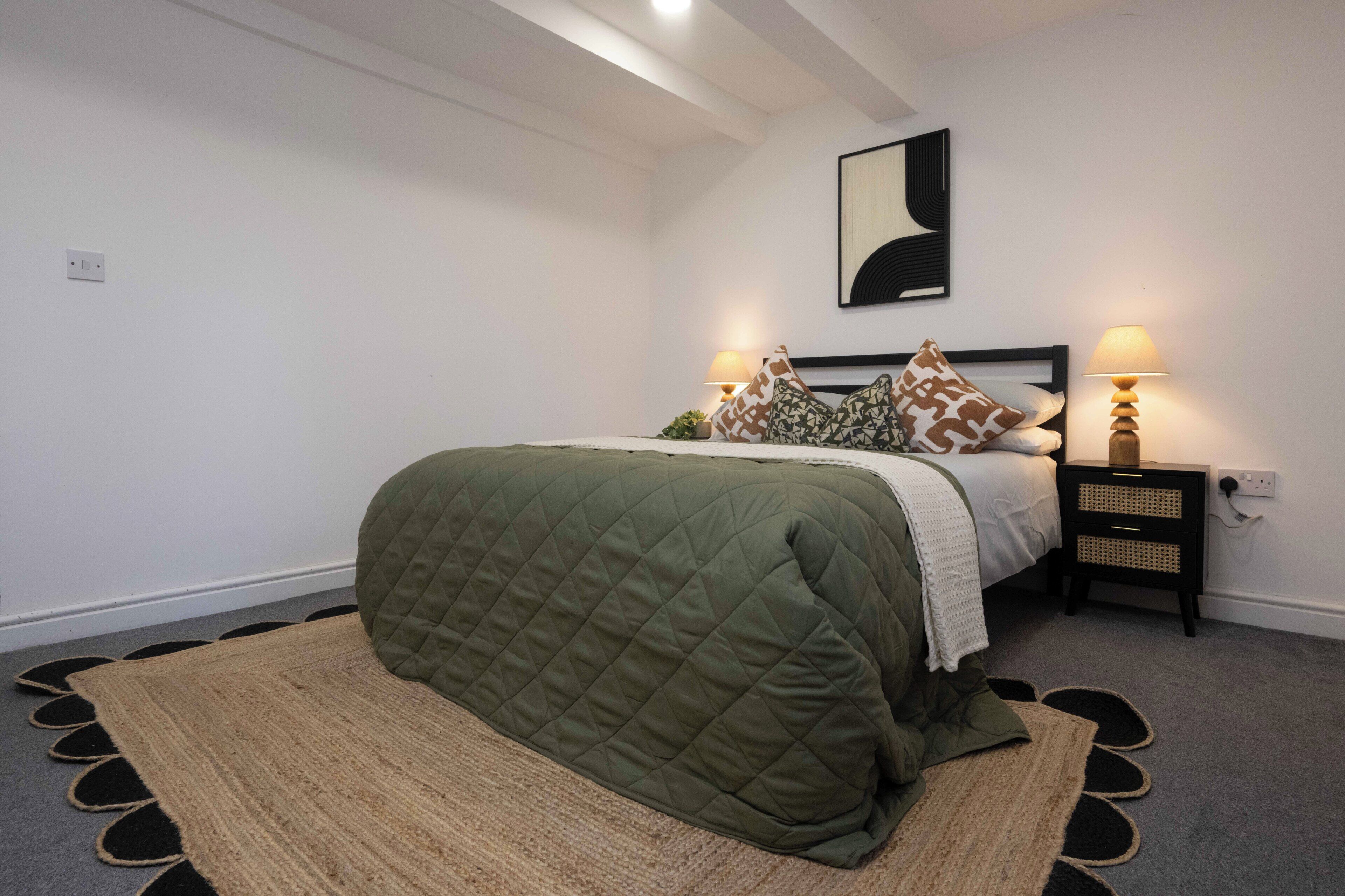 A bedroom with a bed, two bedside lamps, a wall-mounted abstract art piece, and a patterned rug.