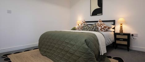 A bedroom with a bed, two bedside lamps, a wall-mounted abstract art piece, and a patterned rug.