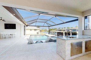 Un espace piscine extérieur moderne doté d’un patio fermé par des baies vitrées, d’une cuisine intégrée avec des meubles en acier inoxydable et d’un coin salon avec des chaises blanches.