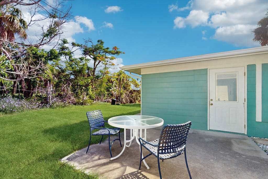 Une maison à l’extérieur vert, avec une porte blanche et un petit patio doté d’une table en verre et de chaises en métal.