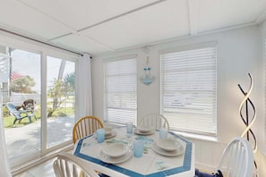 A dining area with a table set for four, chairs, and a view through large windows. - Old Florida bungalow with WiFi, AC, washer/dryer, & patio - walk to beach (Englewood)
