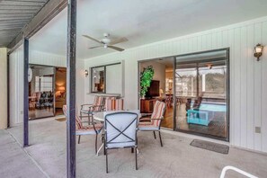 Ein überdachter Patio-Bereich mit Deckenventilator, einem Esstisch mit Stühlen und einer Glasschiebetür, die zu einem Außenbereich führt.