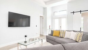 Ein modernes Wohnzimmer mit Flachbildfernseher, Glastisch und grauem Sofa mit gemusterten Kissen.