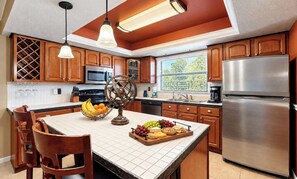 A kitchen with wooden cabinets, a stainless steel refrigerator, a central island with fruit and cheese, and pendant lighting. - Enjoy the serene pool area and back yard! (Cape Coral)