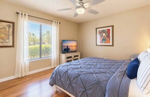 Une chambre à coucher avec un lit, un téléviseur posé sur un meuble, un ventilateur de plafond et une fenêtre avec des stores.