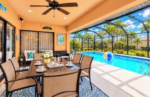 A poolside area with a dining table set for four, a ceiling fan, and a view of a swimming pool with palm trees. - Enjoy the serene pool area and back yard! (Cape Coral)
