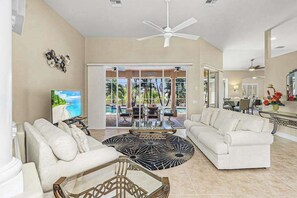 A spacious living room with a large sofa, a coffee table, and a TV. There is a glass door leading to a balcony with a pool. - Exceptional waterfront home, refreshing pool, spa, family and dog friendly (Cape Coral)