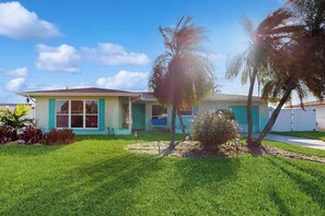 A single-story house with a front porch, a small garden with palm trees, and a clear blue sky. - Waterfront retreat with pool, arcade game, family friendly (Cape Coral)