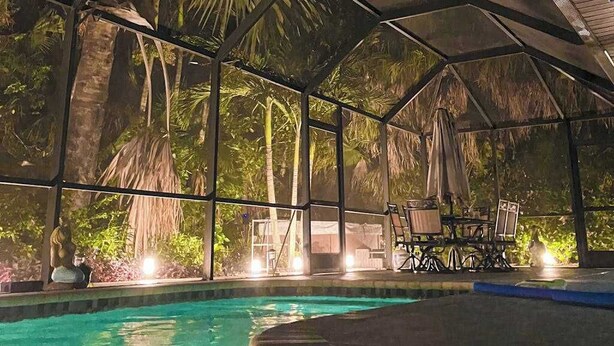Una piscina interior con techo de vidrio, rodeada de plantas tropicales y un área de descanso con mesas y sillas.