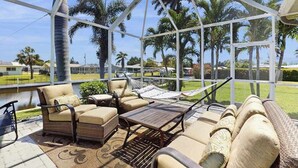 A covered outdoor seating area with wicker furniture, a hammock, and a view of palm trees and a body of water. - Beautiful home with private pool, waterfront views & home theater (Cape Coral)