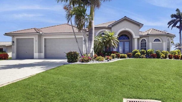 A single-story house with a garage, a well-maintained lawn, and a variety of flowering plants. - Beautiful home with private pool, waterfront views & home theater (Cape Coral)