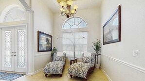 A hallway with two patterned armchairs, a chandelier, a framed picture, and a window with blinds. - Beautiful home with private pool, waterfront views & home theater (Cape Coral)