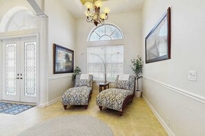 A hallway with two patterned armchairs, a chandelier, a framed picture, and a window with blinds. - Beautiful home with private pool, waterfront views & home theater (Cape Coral)
