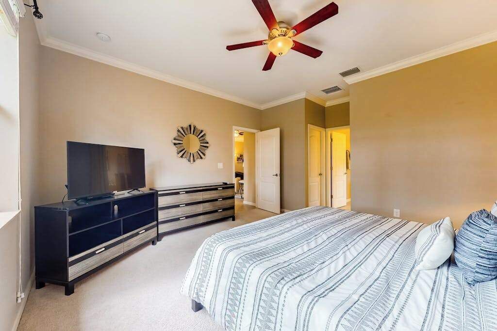 Une chambre à coucher avec un lit, un téléviseur posé sur une commode, un ventilateur de plafond et une porte donnant sur une autre pièce.