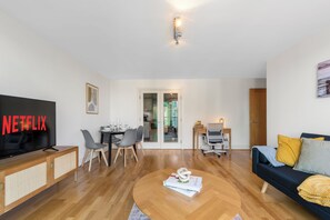 Ein modernes Wohnzimmer mit Flachbildfernseher, einem Essbereich mit Stühlen und Tisch, einer Couch mit Kissen und einem Holztisch.
