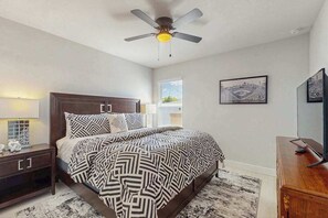 Een slaapkamer met een bed, nachtkastje, dressoir, plafondventilator en een raam met jaloezieën.