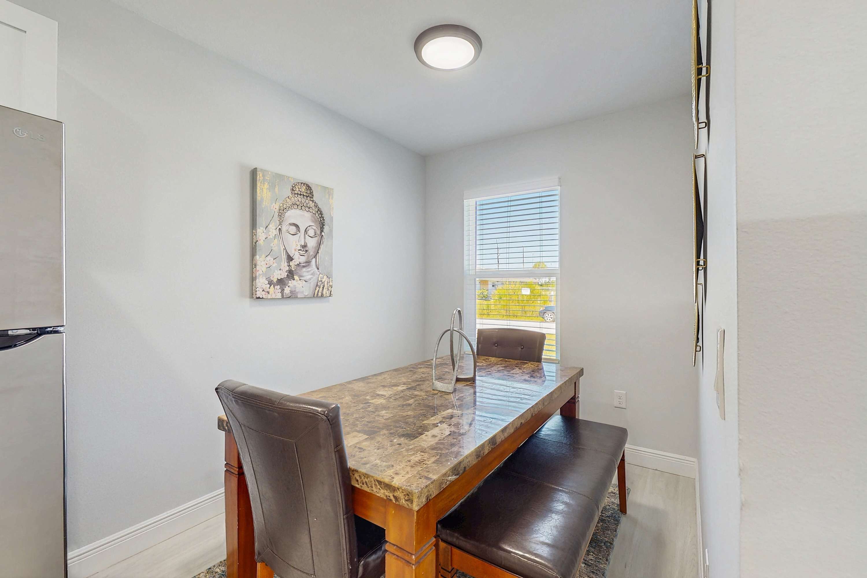 A dining area with a marble table, leather chairs, a bench, a Buddha painting, and a window with blinds.