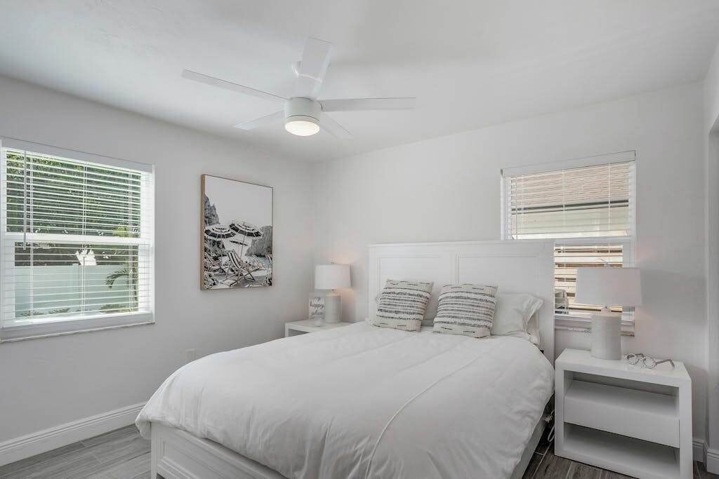 Un dormitorio blanco con cama, mesitas de noche, ventilador de techo y ventana con persianas.