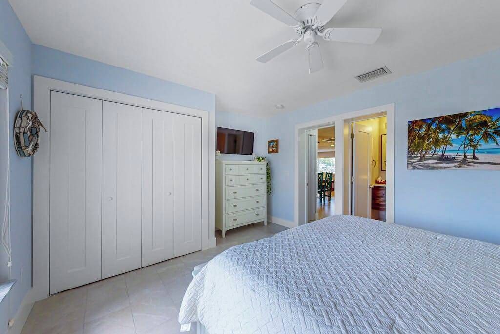 A bedroom with a bed, a white wardrobe, a dresser, a ceiling fan, and a painting of a beach scene.
