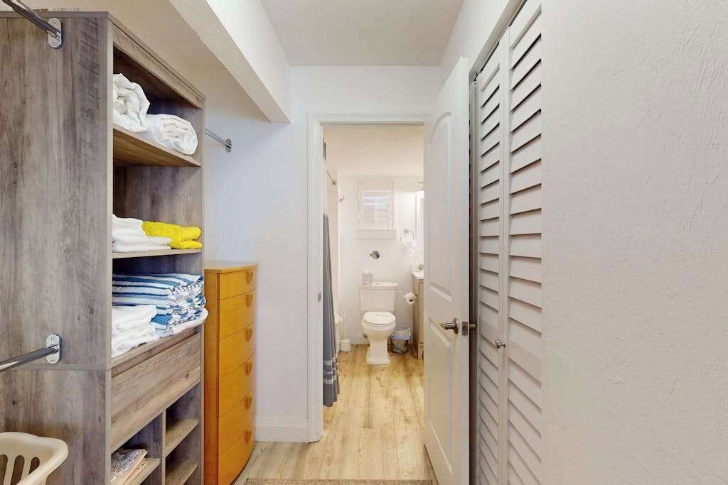 A narrow hallway leading to a bathroom with a toilet, a wooden cabinet with folded towels, and a door with horizontal slats.