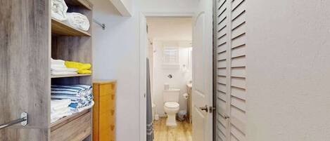A narrow hallway leading to a bathroom with a toilet, a wooden cabinet with folded towels, and a door with horizontal slats.