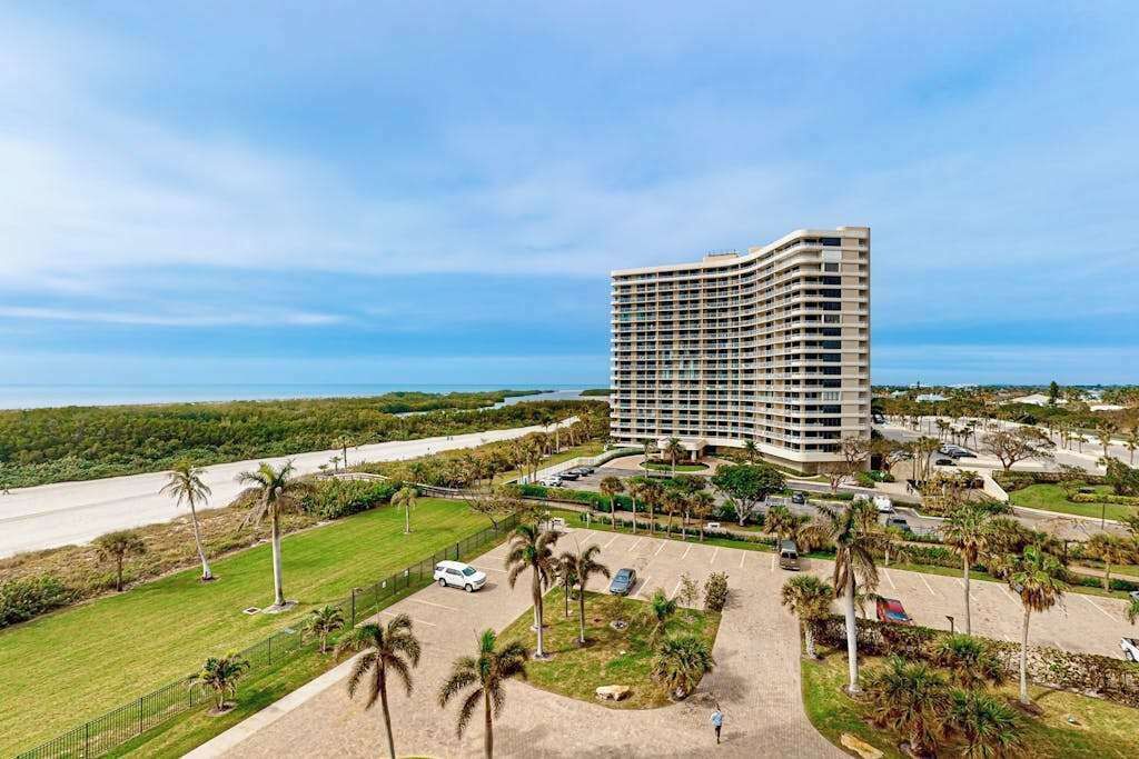 A high-rise hotel with a parking lot, palm trees, and a view of the ocean.