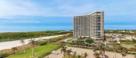 A high-rise hotel with a parking lot, palm trees, and a view of the ocean.