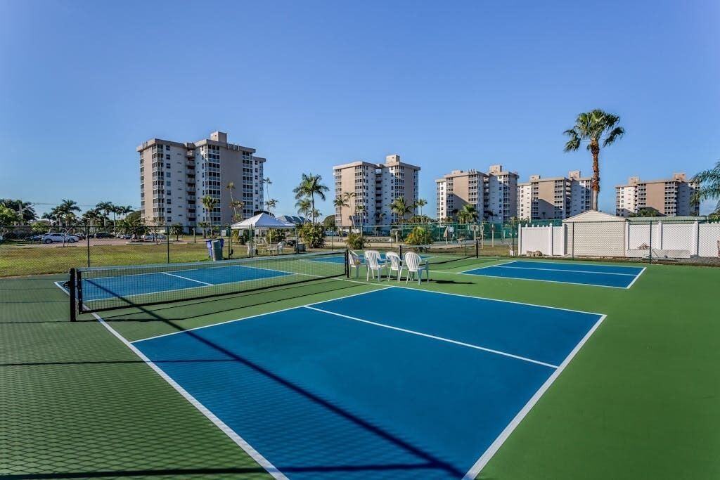 A tennis court with a net, surrounded by a fence, and palm trees in the background.