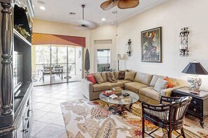 A living room with a sectional sofa, a round coffee table, a patterned rug, a dining area with a table and chairs, and a large window with a view. - Gated-entry townhome with on-site golf, pool, gym, clubhouse & rec amenities (Bonita Springs)