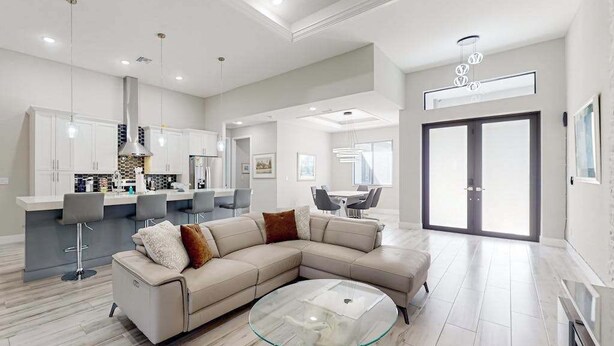 A modern living room with a sectional sofa, a glass coffee table, and a kitchen area with bar stools. - Luxurious home with private washer/dryer, enclosed yard, WiFi, & screened lanai (Bonita Springs)