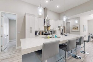 A modern kitchen with white cabinets, a large island with bar stools, pendant lighting, and a stainless steel range hood.