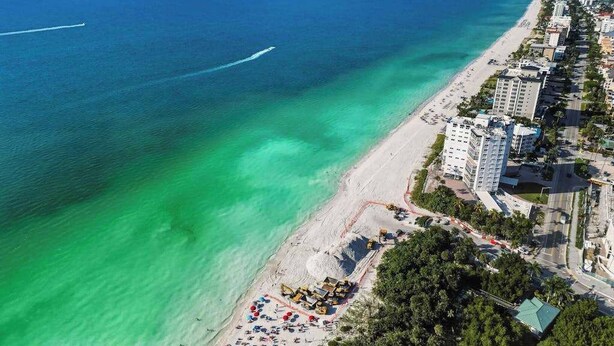 Vista aérea de una zona costera con playa, edificios y mar azul transparente.