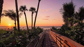 Un sendero de madera que conduce a una playa con palmeras y un atardecer.
