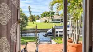 A patio with a chair, a view of a house across a water body, and a large potted plant. - Villa Euphoria: Chic 2-unit, canal-front retreat with 2 pools & private dock (Cape Coral)