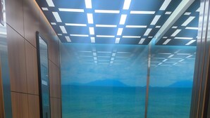 En elevator med udsigt til en træterrasse, der fører ned til et vandareal, med et bjerg i det fjerne.
