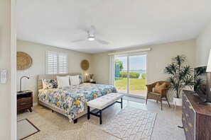 Ein Schlafzimmer mit einem Bett, einer Bank, einem Nachttisch, einem Schminktisch, einem Deckenventilator, einem Fenster und Blick ins Freie.