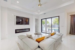 A modern living room with a sectional sofa, a flat-screen TV, a fireplace, and a large sliding glass door leading to an outdoor area. - New modern home with pool, lawn, W/D, Wifi and AC (Cape Coral)