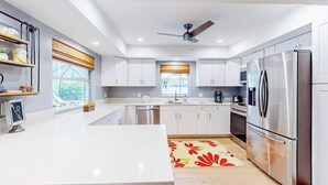 Una cocina moderna con armarios blancos, electrodomésticos de acero inoxidable, un ventilador de techo y una ventana con persianas.