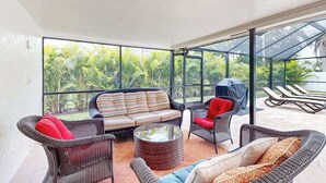 Un espacio de estar al aire libre techado con mobiliario de mimbre y una mesa de vidrio, con vistas a un exuberante jardín.