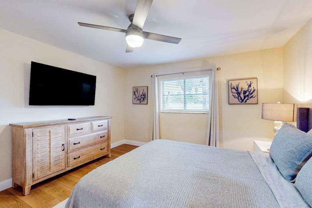 A bedroom with a bed, a ceiling fan, a TV, a dresser, and a window with blinds.