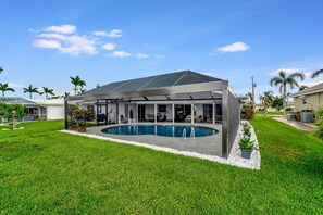 A covered outdoor pool area with a modern architectural design, surrounded by a well-maintained lawn and palm trees. - Dog-friendly water-view house with pool & firepit - snowbirds welcome (Cape Coral)