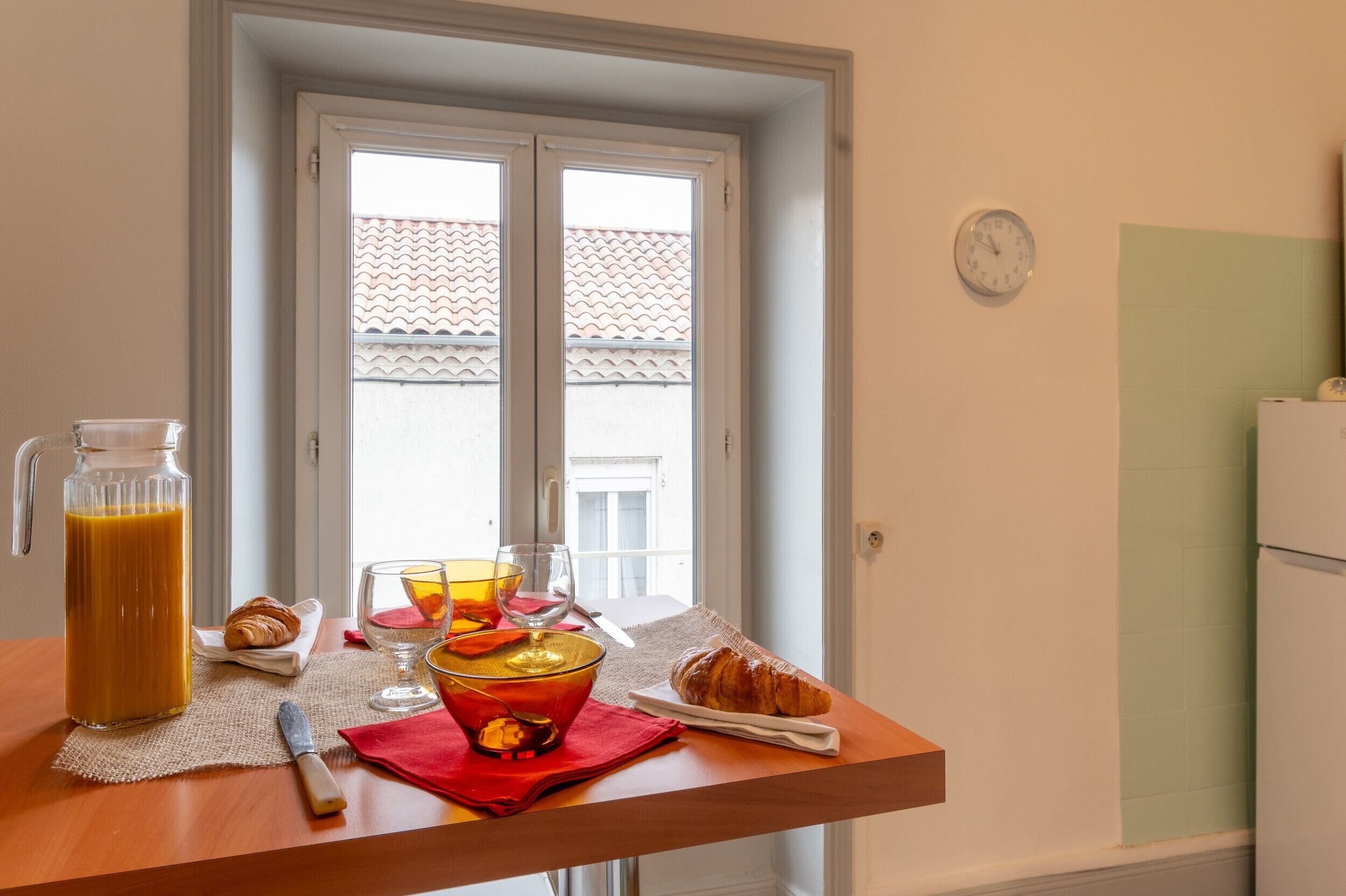 Una mesa junto a una ventana con zumo de naranja en una jarrón, cruasanes y un vaso de zumo de naranja.