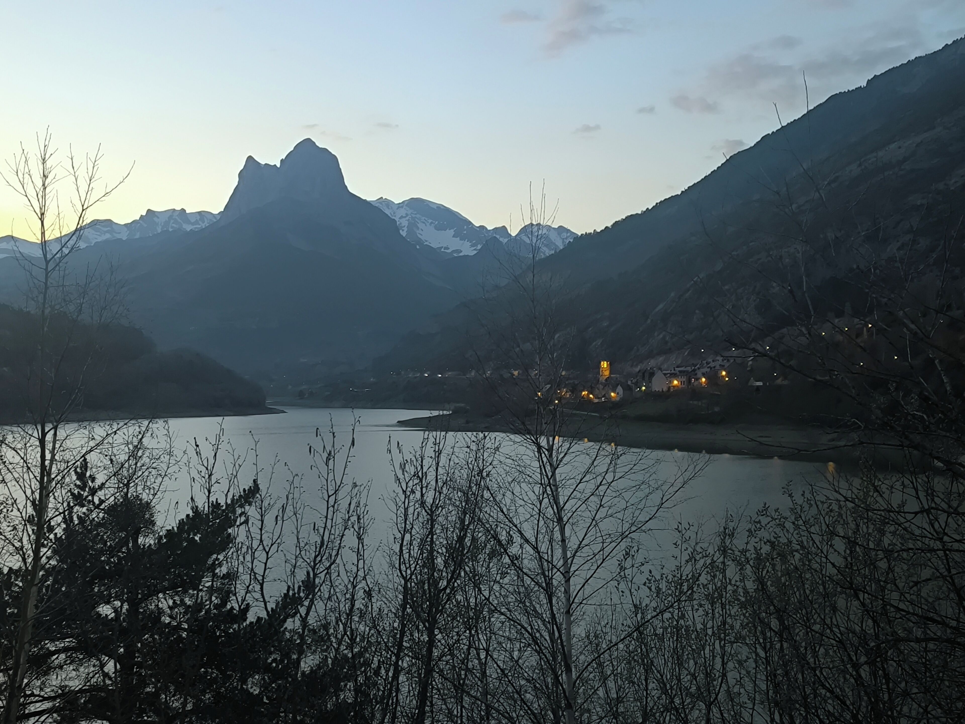 Paisaje montañoso con un lago, árboles y un pequeño asentamiento con luces.
