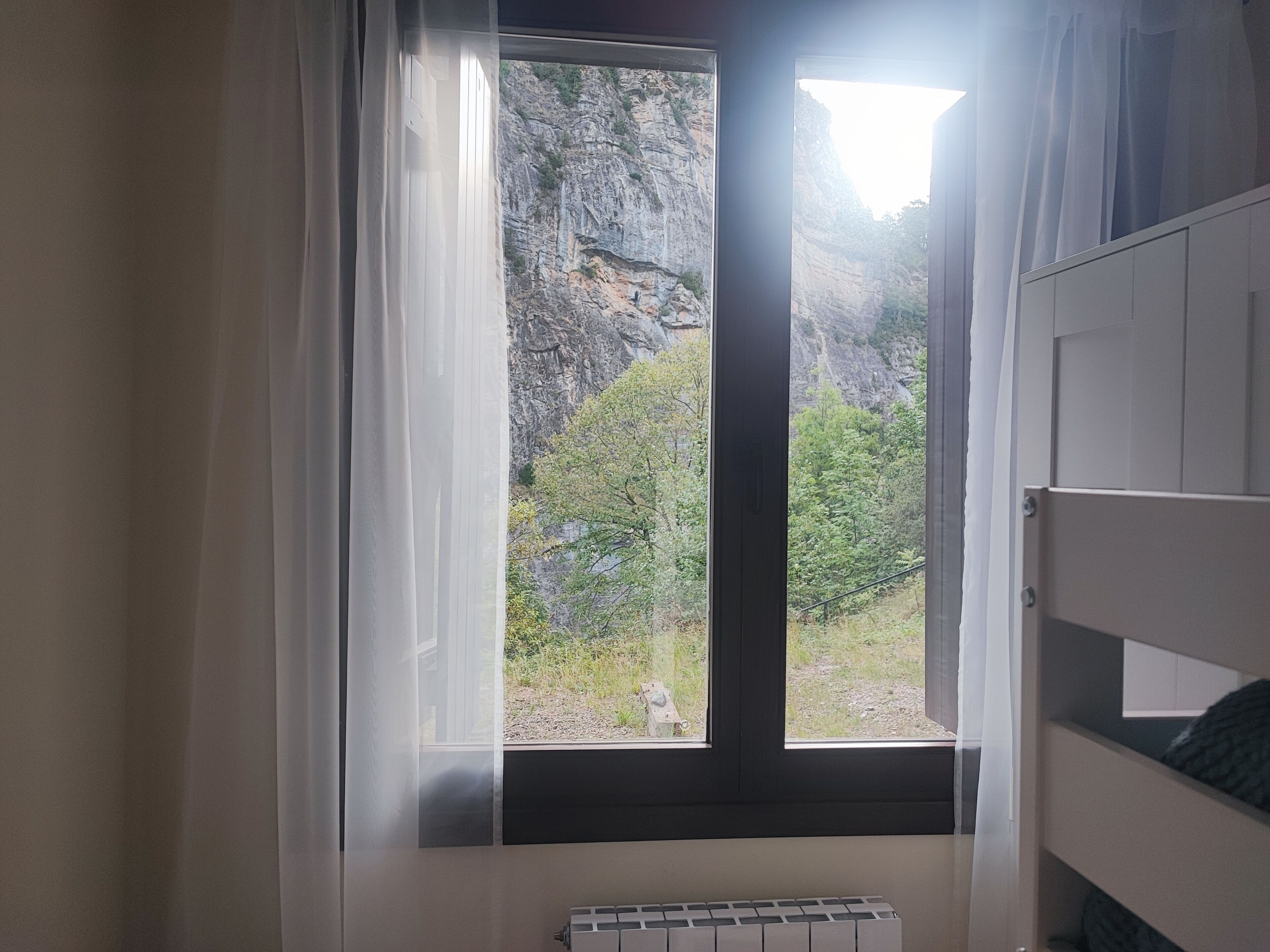 Una habitación con ventana a un acantilado rocoso y vegetación, un radiador y una cama litera.