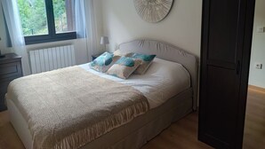 Ein Schlafzimmer mit Bett, Nachttisch, Lampe, Fenster mit Vorhängen, Heizkörper, Wanddekoration und Kleiderschrank.