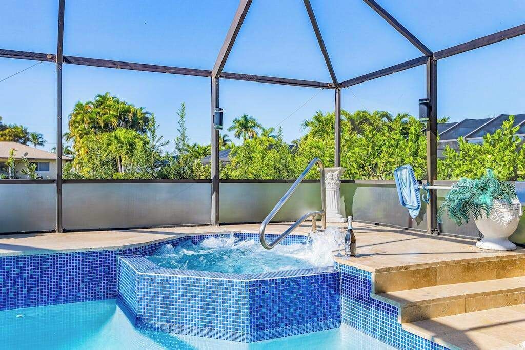 A hot tub with a metal frame and a tiled floor, surrounded by a covered area with a view of palm trees and a building in the background.