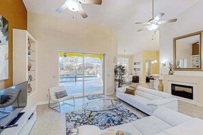 A spacious living room with a large window, a white sofa, a glass coffee table, and a ceiling fan. - Canal-front home with pool, outdoor kitchen, & private dock - dog-friendly (Cape Coral)