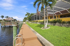A waterfront walkway with a wooden deck, palm trees, and a building with a glass facade. - Waterfront Hideaway | Pool & Private Dock (Cape Coral)