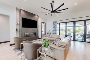 A modern living room with a fireplace, a large TV, a ceiling fan, and a view of the outdoors. - Luxury canal front home with heated pool, pool spa, grill, fireplace, W/D, & AC (Cape Coral)
