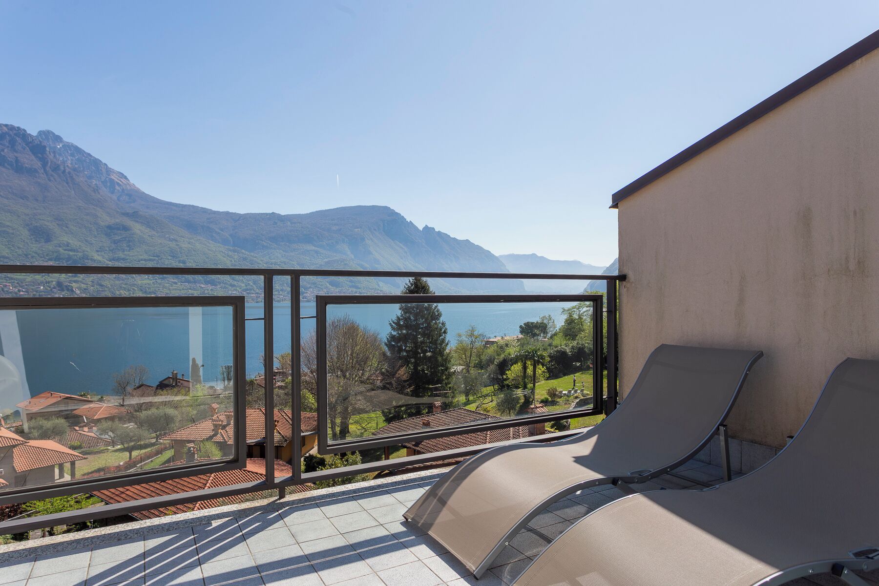 A balcony with a view of a lake and mountains, two lounge chairs, and a glass railing.